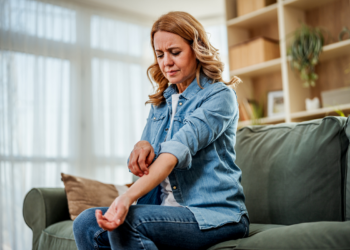 Woman sitting on a couch scratching her itchy forearm, wincing in discomfort