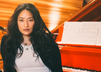 Chloe Flower seated in front of a red grand piano with sheet music, wearing a black jacket over a white top.