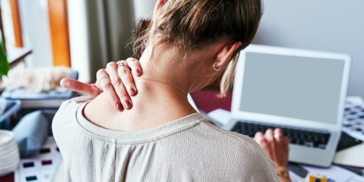 Woman seen from behind at a laptop, hand pressed to the back of her neck as she tilts her head forward.